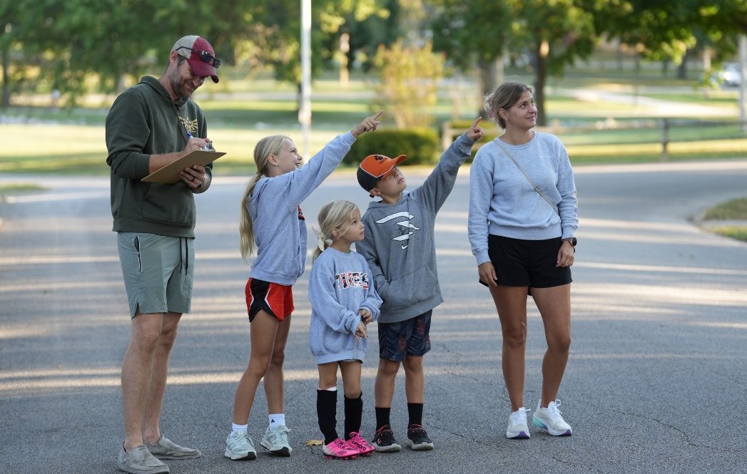 An Olney family on a residential street pointing up at a squirrel in the trees, one of them writing on a clipboard